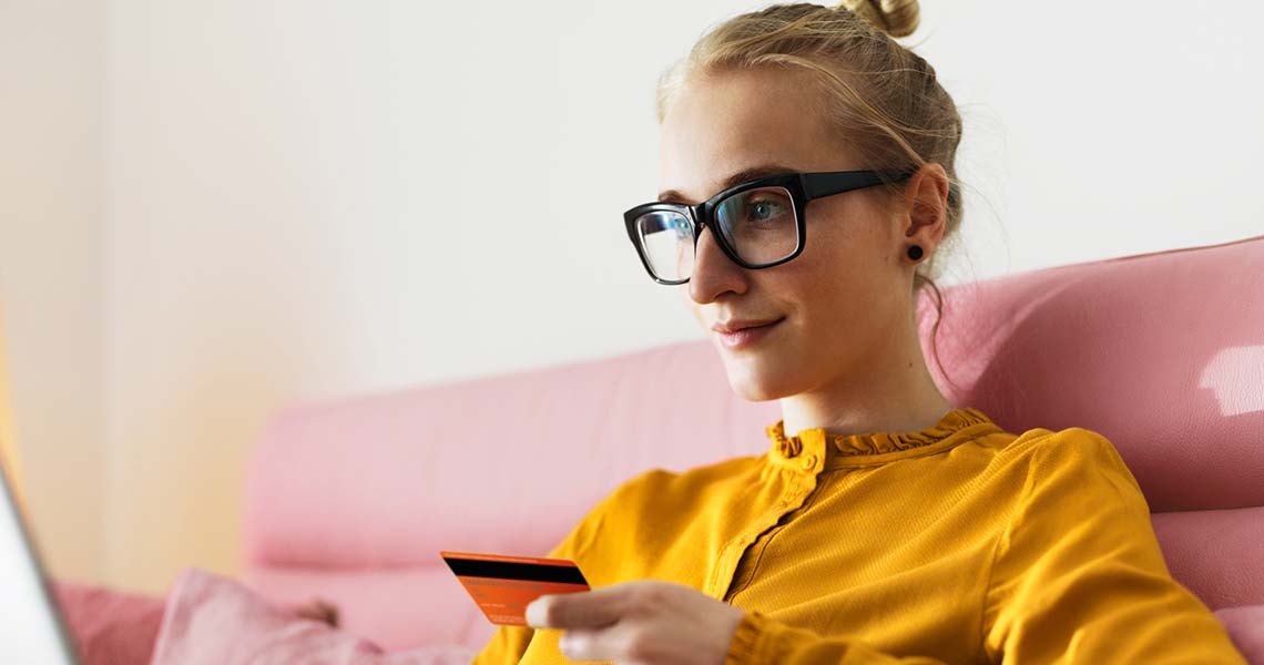 Jente i briller og gul genser som sitter i en rosa sofa med betalingskort og bærbar PC